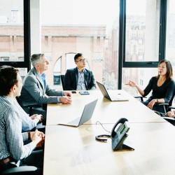 Mehrere Mitarbeiter sitzen in einem modernen Büro an einem Konferenztisch und diskutieren, im Hintergrund eine große Fensterfront, durch die man ein modernes Industriegebäude aus rotem Backstein sieht