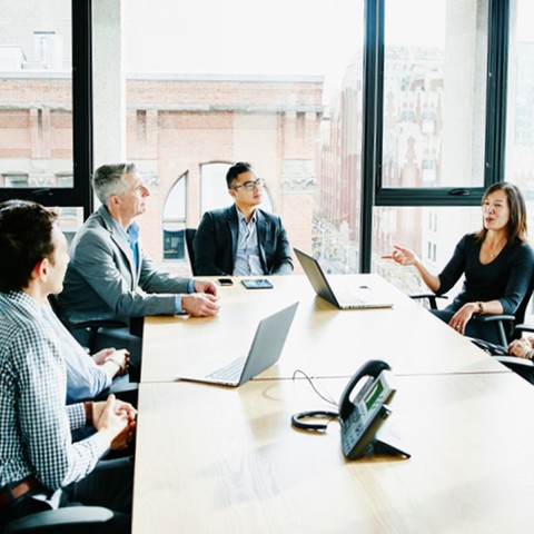 Mehrere Mitarbeiter sitzen in einem modernen Büro an einem Konferenztisch und diskutieren, im Hintergrund eine große Fensterfront, durch die man ein modernes Industriegebäude aus rotem Backstein sieht