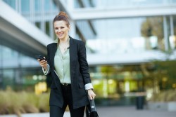 Junge Frau im Anzug in die Kamera lachend draussen mit Handy und Tasche vor Bürogebäude
