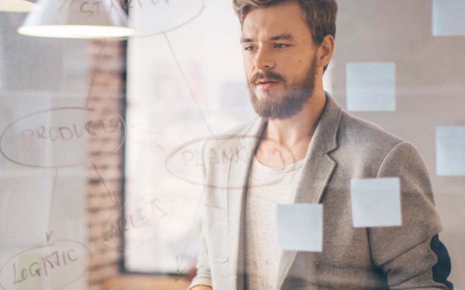 Dunkelhaariger Mann mit Bart steht in einem Büro vor einem Flipchart und schaut nachdenklich darauf