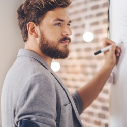 Dunkelhaariger Mann mit Bart steht in einem Büro vor einem Flipchart und zeichnet eine Art Mmindmap darauf