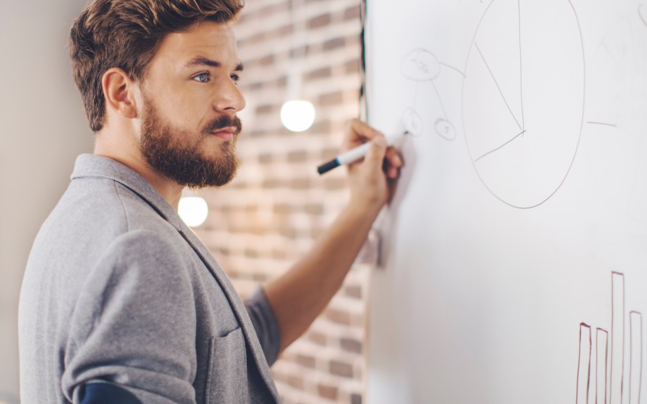 Dunkelhaariger Mann mit Bart steht in einem Büro vor einem Flipchart und zeichnet eine Art Mmindmap darauf
