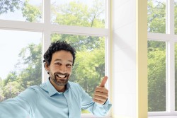 Ein Mann steht lächelnd vor einem Fenster und hält ein Daumen hoch in die Kamera