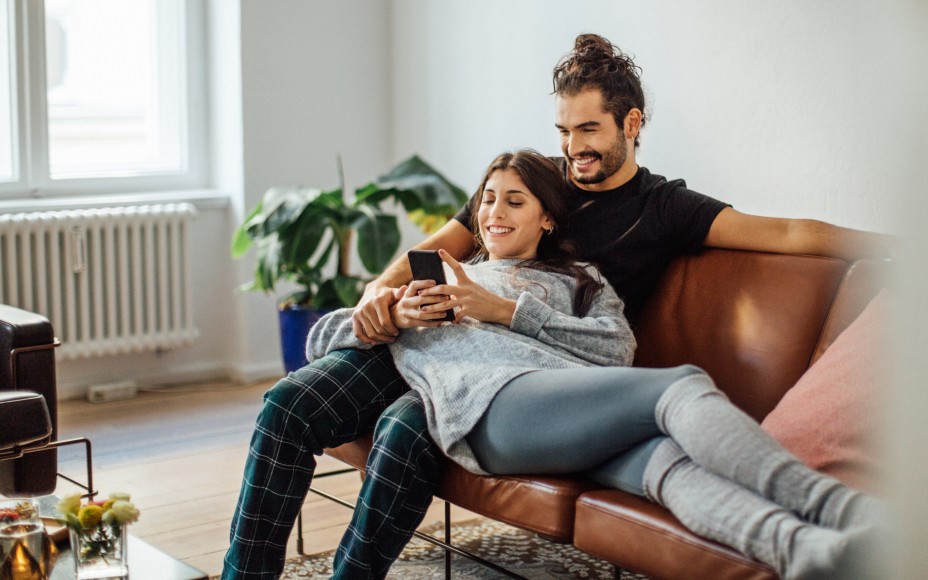Junges Paar kuschelt auf dem Sofa während sie gemeinsam auf das Smartphone der Frau schauen