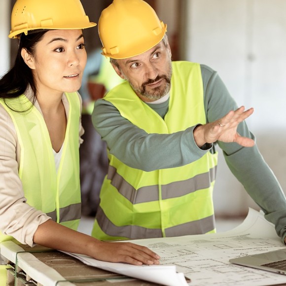 Frau und Mann in Schutzhelm und Warnweste besprechen einen Bauplan auf einer Baustelle