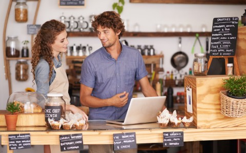 Die Ladenbesitzer eines Cafes, ein junges Paar, schauen sich lächelnd an, während er vor einem geöffneten Laptop auf der der Theke steht. Im Vordergrund sind Muffins zu sehen, im Hintgergrund Kaffeekannen