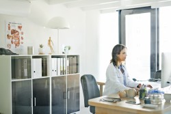 Junge Ärztin im weißen Kittel sitzt an ihrem hölzernen Schreibtisch vor dem Computer und tippt etwas. Im Hintergrund ein weißes Regal mit Akten, darüber ein Poster mit menschlichen Organen an der Wand