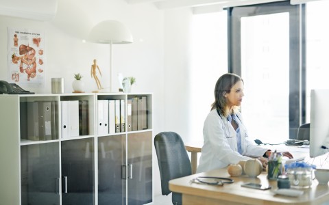 Junge Ärztin im weißen Kittel sitzt an ihrem hölzernen Schreibtisch vor dem Computer und tippt etwas. Im Hintergrund ein weißes Regal mit Akten, darüber ein Poster mit menschlichen Organen an der Wand
