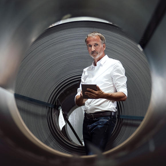 Mann mit weißem Hemd und grauem Bart steht mit einem Tablet in der Hand vor einer schwarzen Röhre