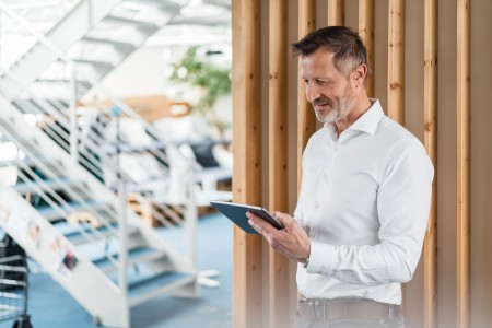 Junger Mann in weißem Hemd lehtn mit einem Laptop an einer Wand mit Holzpaneelen, im Hintergrund führt eine offene Treppe mit Glas in eine obere Etage