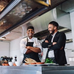 Gastronom im Anzug und Koch mit Schürze und Kopfbedeckung stehen lachend mit verschränkten Armen in einer modernen Restaurantküche