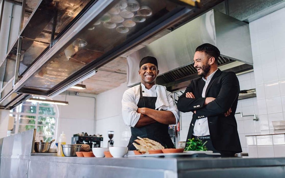 Gastronom im Anzug und Koch mit Schürze und Kopfbedeckung stehen lachend mit verschränkten Armen in einer modernen Restaurantküche