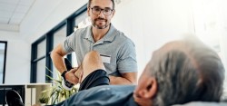 Physiotherapeut mit Brille im grauen Polo-Shirt dehnt das Bein eine Mannes in kurzer Sporthose und Sportshuhen und knickt es im Kniegelenk