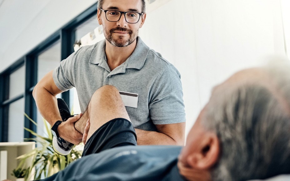 Physiotherapeut mit Brille im grauen Polo-Shirt dehnt das Bein eine Mannes in kurzer Sporthose und Sportshuhen und knickt es im Kniegelenk
