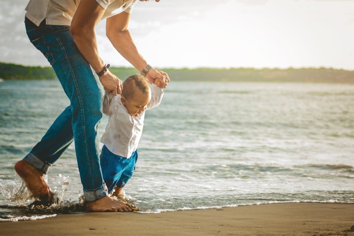 Vater hält sein kleines Kind an beiden Händen und unterstützt es beim Laufen lernen über einen Strand, beide sind barfuß