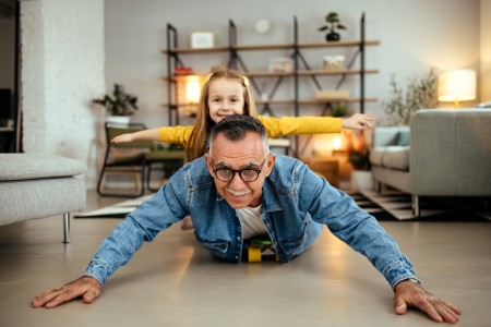 Vater liegt auf dem Bauch im Wohnzimmer mit seiner Tochter, beide haben die Arme seitlich ausgestreckt und lachen in die Kamera