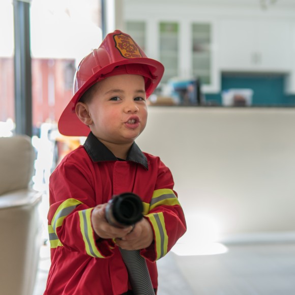 Kleiner Junge, der als roter Feuerwehrmann verkleidet ist, steht im Wohnzimmer und hält einen Staubsaugerschlauch und tut so, als ob er einen Brand löscht