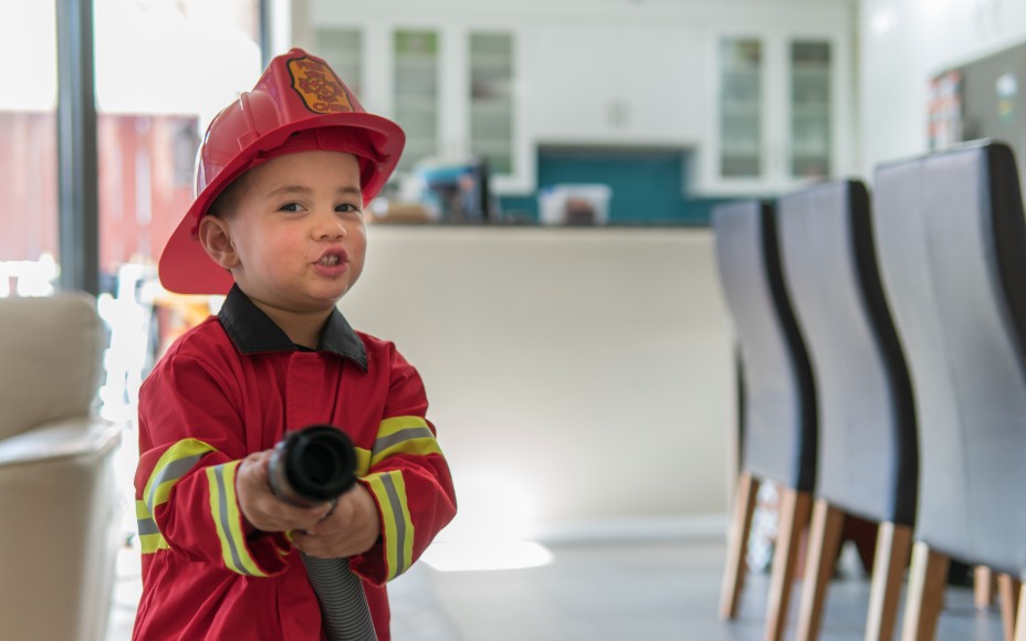 Kleiner Junge, der als roter Feuerwehrmann verkleidet ist, steht im Wohnzimmer und hält einen Staubsaugerschlauch und tut so, als ob er einen Brand löscht