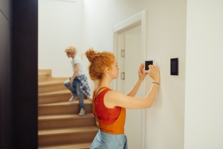 Frau mit roten, hochgesteckten Haaren im orangefarbenen Shirt programmiert eine Alarmanlage an der Wand, während im Hintergrund ein Kind eine moderne Holztreppe nach oben geht