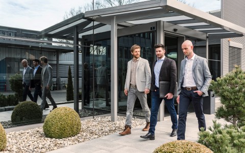 Drei Geschäftsleute in legerer Kleidung gehen nebeneinander an einem einstöckigen Bürogebäude mit vielGlas vorbei