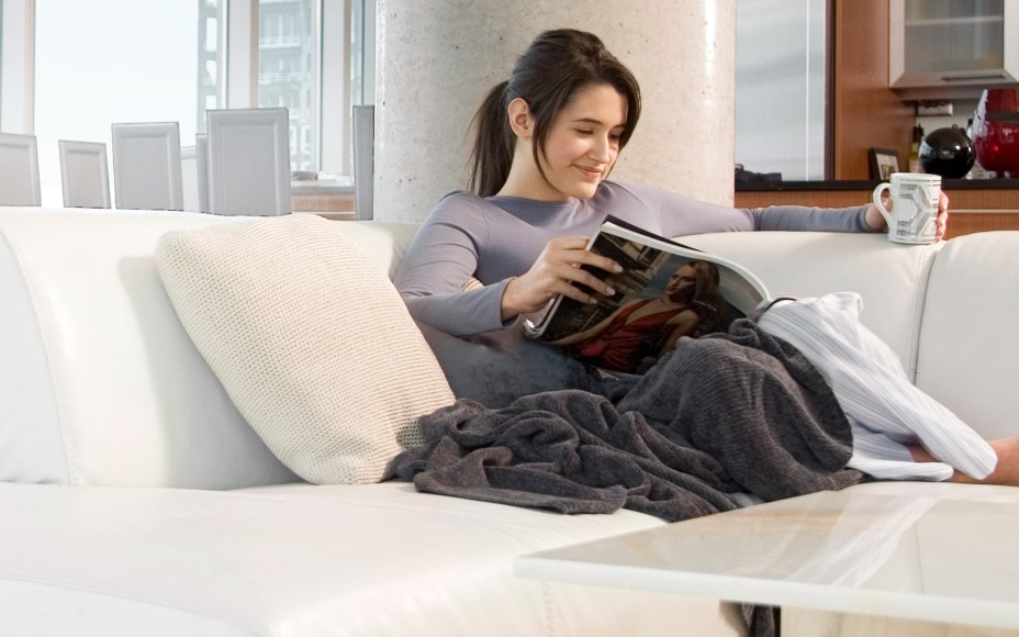 Dunkelhaarige Frau sitzt auf einem hellen modernen Sofa in eine Decke gekuschelt mit einer tasse in der linken Hand und liest in einer Zeitschrift