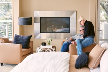 Wohnlandschaft mit lachsfarbenen Ledersofas und großem Flatscreen auf der Kopfwand, rechts im Bild sitzt ein älterer Mann in einem passenden Sessel, seine Partnerin sitzt auf der Armlehne und beide schauen etwas auf dem Tablet
