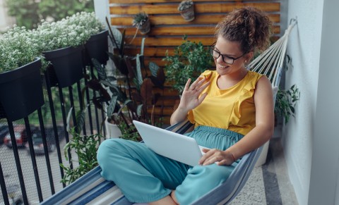 Junge Frau liegt mit gekreuzten Beinen in einer Hängematte auf einem begrünten Balkon und winkt in ihr Tablet.