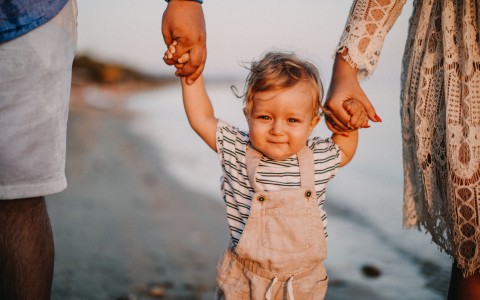 Eltern gehen mit ihrem kleinen Kind am Meer in der Abendsonne spazieren, sie halten das Kind in der Mitte von sich beide an den Händen, man sieht von ihnen nur die Beine
