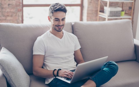 Junger Mann mit Bart sitzt lächelnd im Schneidersitz auf einem cremefarbenen Sofa und tippe auf seinem Laptop