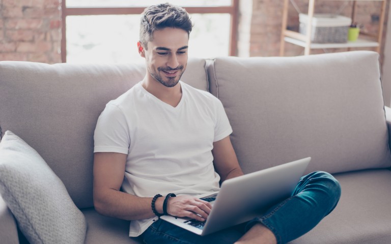 Junger Mann mit Bart sitzt lächelnd im Schneidersitz auf einem cremefarbenen Sofa und tippe auf seinem Laptop