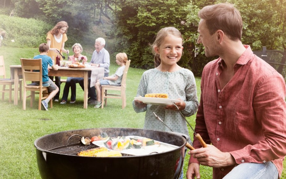 Familie mit drei Kindern grillt mit den Großeltern im Garten, Vater und Tochter stehen am Holzkohlegrill und lachen sich an, Rest der Familie sitzt weiter hinten im Bild am Tisch, ganz hinten ist ein Foliengewächshaus zu sehen