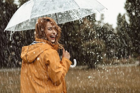 Junge Frau im gelben Regenmantel trägt einen durchsichtigen Regenschirm, geht im starken Regen spazieren und schaut lachend nach hinten über die rechte Schulter in die Kamera
