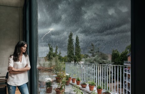 Frau lehnt an einer großen Fensterfront und schaut hinaus in den Garten während draußen ein Gewitter tobt, der Himmel dunkelgrau und voller Blitze ist