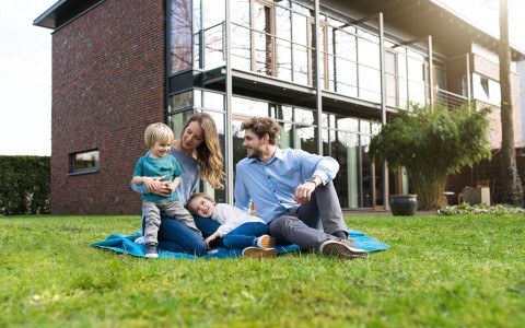 Eine glückliche Familie sitzt mit kleinem blonden Sohn im Garten vor ihrem Haus auf einer Decke.