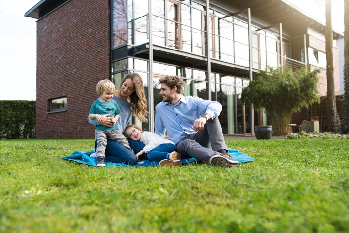 Eine glückliche Familie sitzt mit kleinem blonden Sohn im Garten vor ihrem Haus auf einer Decke.