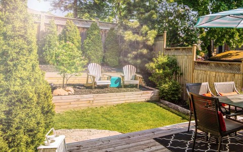 Moderner Garten von einem Holzzaun umgeben. links ein paar große Konifären, rechts eine Sitzgruppe für 6 Personen mit roten Kissen unter einem grün-weiß-gestreiften Sonnenschirm