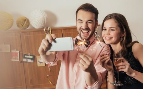 Junges Paar macht lachend ein Selfie, er hält brennende Wunderkerzen in der Hand ud sie ein Sektglas