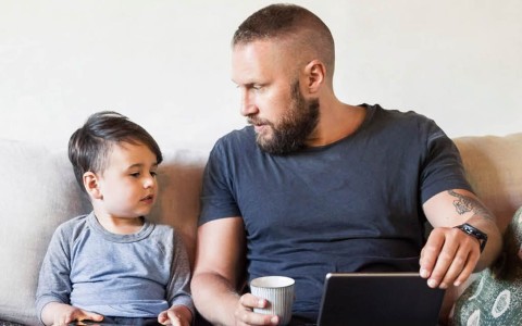 Vater und kleiner Sohn, ein Kleinkind, sitzen nebeneinander auf einem hellen Sofa mit bestickten Kissen und sehen sich an, der Vater hat ein Tablet auf dem Schoß und eine Tasse in der Hand