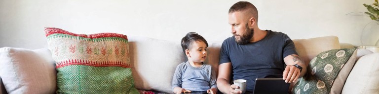 Vater und kleiner Sohn, ein Kleinkind, sitzen nebeneinander auf einem hellen Sofa mit bestickten Kissen und sehen sich an, der Vater hat ein Tablet auf dem Schoß und eine Tasse in der Hand