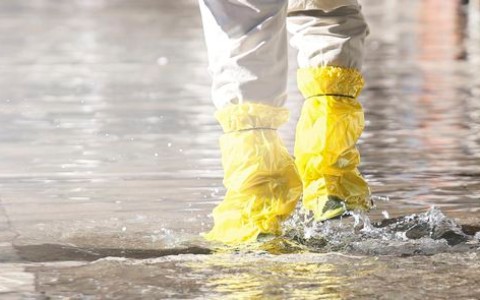 Zwei Menschen, von denen man nur die mit gelbem Wasserschutz versehenen Unterschenkel sieht, waten über eine überflutete Straße
