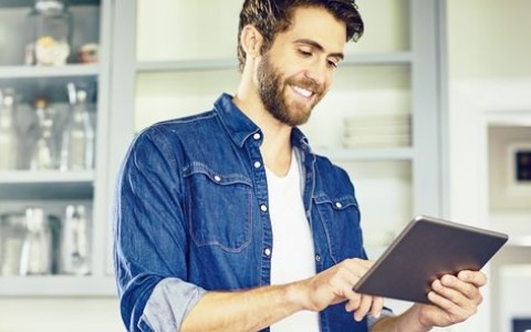 Mann im blauen Jeanshemd tippt etwas auf seinem Laptop während er in seiner Küche steht, im Hintergrund sieht man Küchen-Hängeschränke mit Milchglastüren
