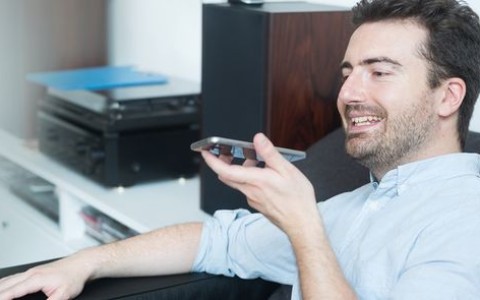 Mann im Hemd sitzt im Wohnzimmer neben seiner Stereoanlage mit Lautsprechern und spricht etwas sein Handy
