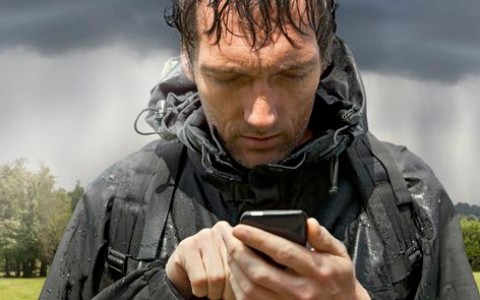 Mann mit nassen Haaren in einem schwarzen Parka und Rücksack steht auf einer Wiese und tippt in sein Handy, im Hintergrund ist ein Wald zu sehen und man sieht den Mann frontal