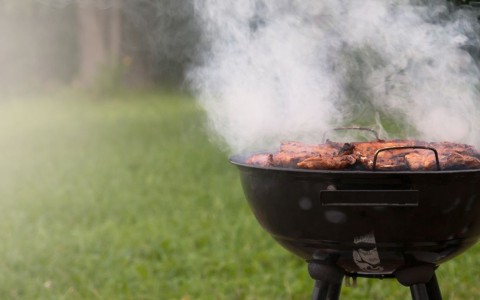 Qualmender schwarzer Holzkohkegrill steht auf einer Wiese und es liegen fast fertige Steaks darauf