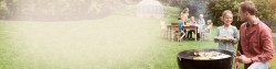 Familie mit drei Kindern grillt mit den Großeltern im Garten, Vater und Tochter stehen am Holzkohlegrill und lachen sich an, Rest der Familie sitzt weiter hinten im Bild am Tisch, ganz hinten ist ein Foliengewächshaus zu sehen