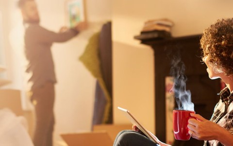 Frau mit kurzen Locken sitzt mit angezogenen Beinen, einer dampfenden roten Tasse und einem Tablet an der wand und schaut zu ihrem Mann, der ein Bild aufhängt