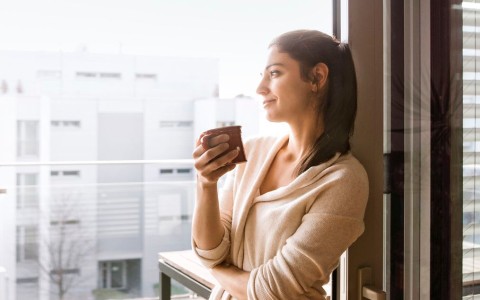 Junge Frau lehnt mit einer Tasse in der Hand am Fensterrahmen in einem höher gelegenen Wohnung und schaut aus dem Fenster mit Blick auf ein modernen Mehrfamilienhaus.