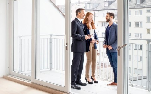 Immobiliernmakler steht mit einem jungen Paar auf dem Balkon einer leehrstehdenen Wohnung in einer höher gelegenen Etage. Alle sind im Businesslook gekleidet.