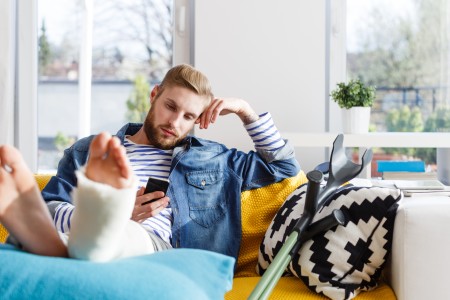 Blonder Mann mit Bart schaut etwas genervt auf sein Handy, weil sein Bein gebrochen und eingegipst ist.