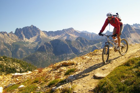 Mountainbiker in roter Jacke, kurzer Hose, Helm und Sonnenbrille bikt auf einem steinigen Trail hoch oben in den bergen, während die oOnne scheint
